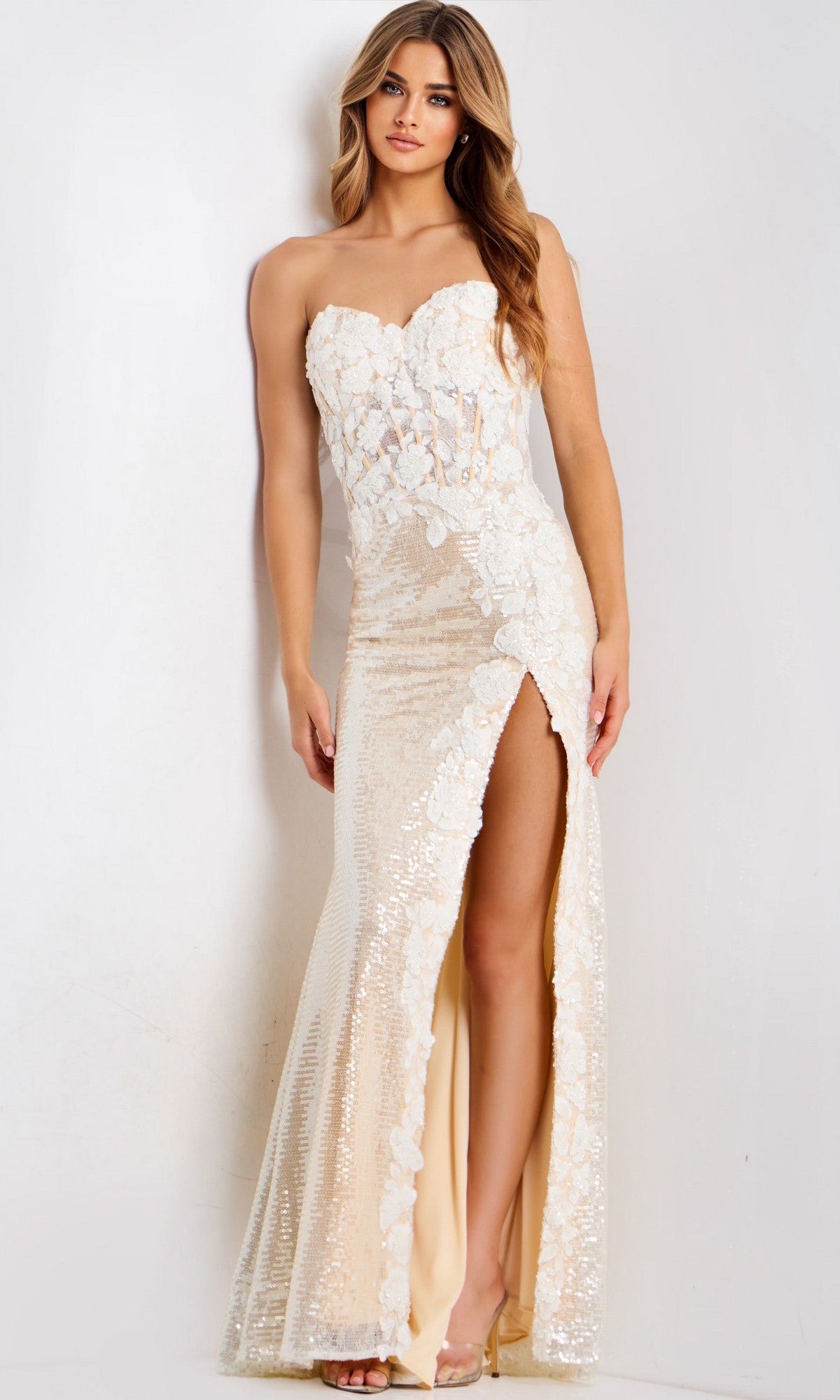 A woman stands against a plain background wearing the Jovani 49362, a strapless, floor-length beige prom dress with floral lace details and a high slit.